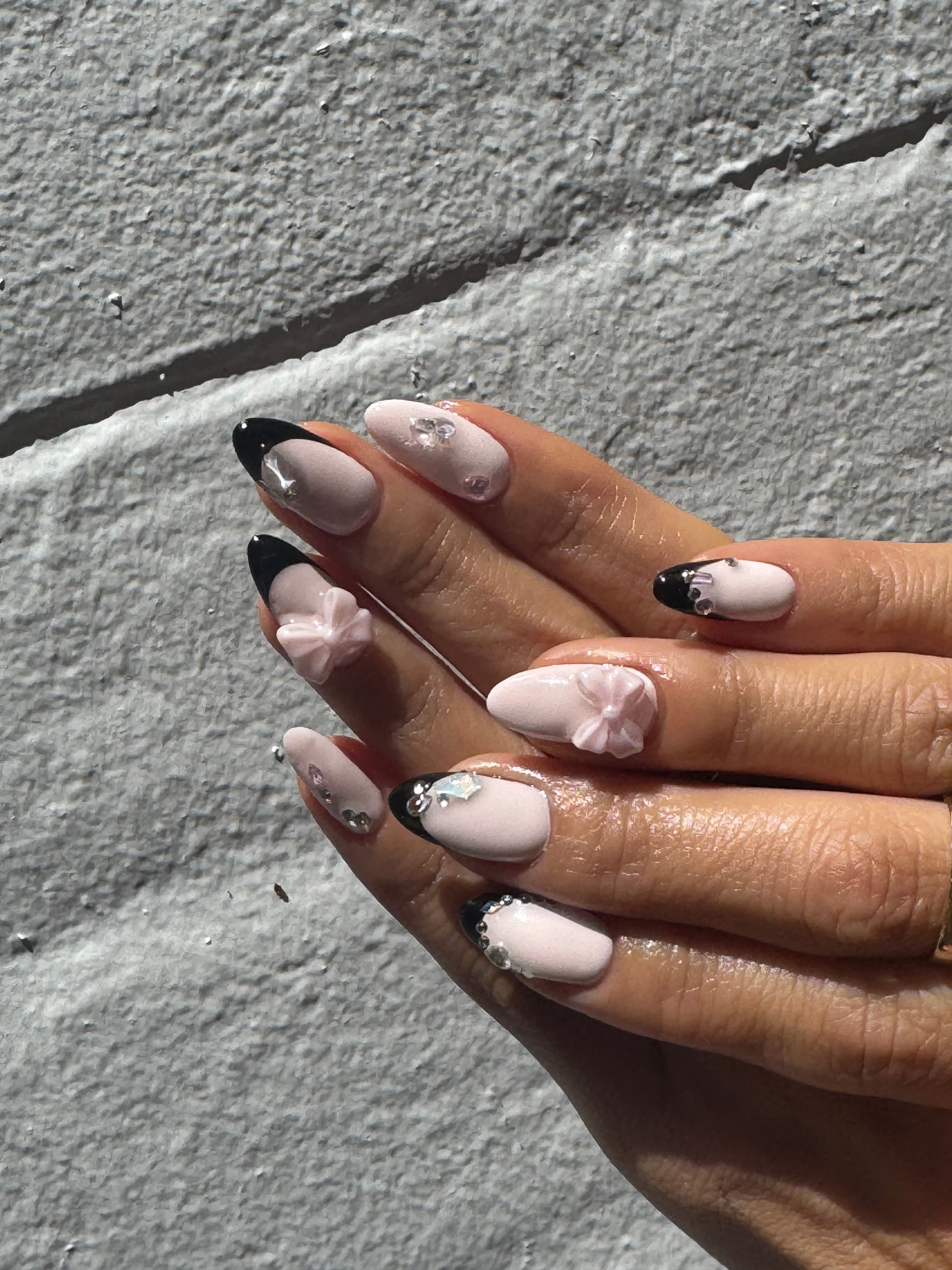 Blush-toned nails with black French tips, bows, and crystal embellishment